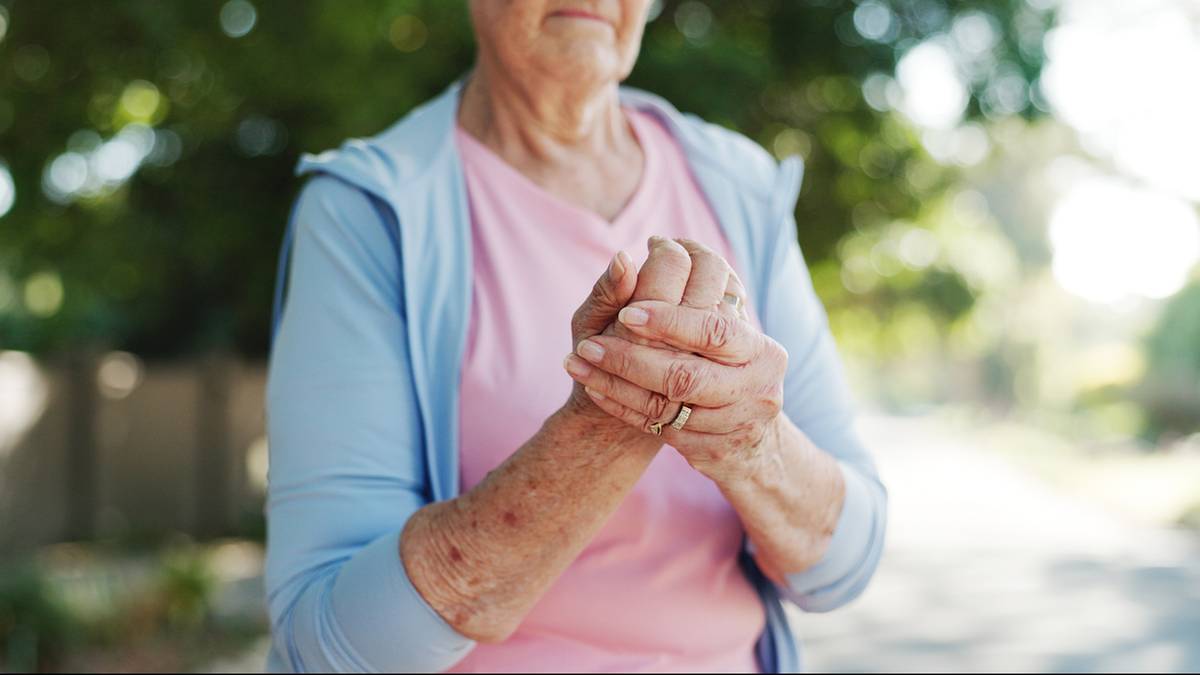 Starsza kobieta trzymająca się za dłonie, symbolizująca potencjalny b&oacute;l staw&oacute;w lub potrzebę siły mięśniowej.