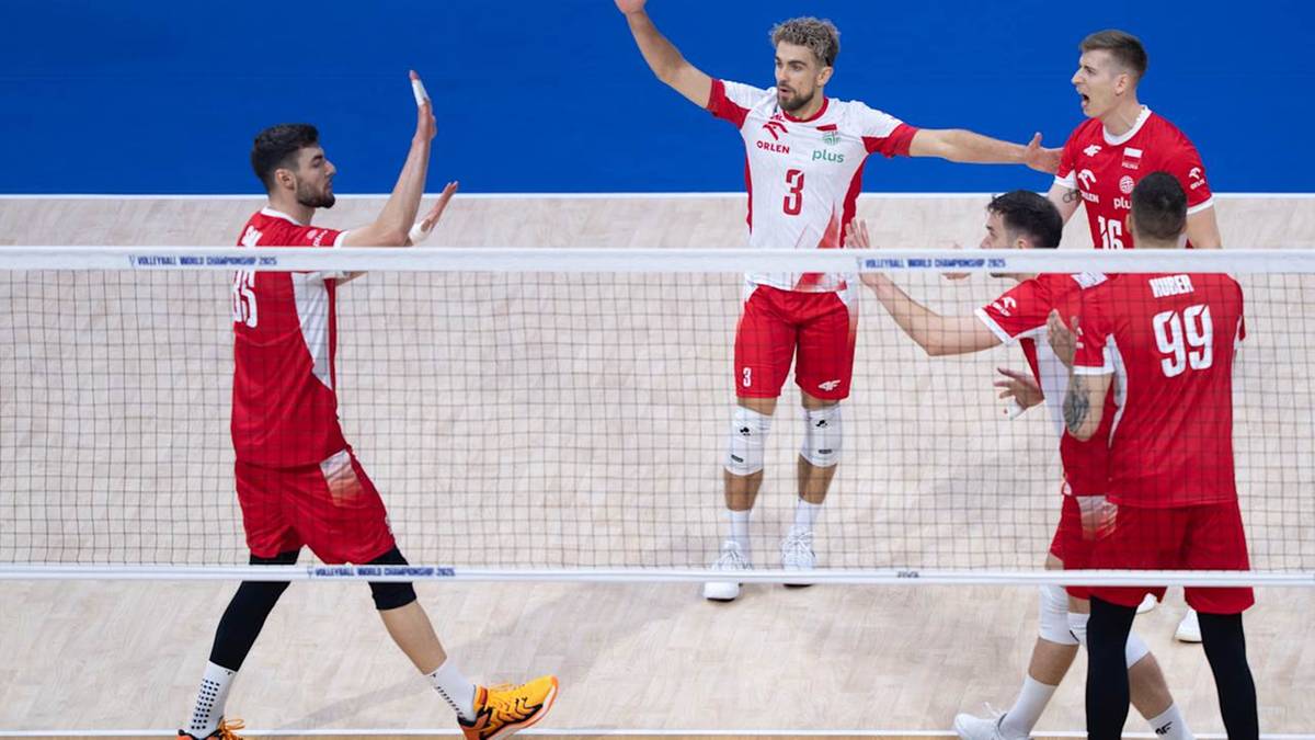 Siatkarze w czerwonych strojach z białymi elementami rywalizują przy siatce. Jeden z graczy unosi ręce w geście radości, podczas gdy jego koledzy z drużyny świętują zwycięstwo.