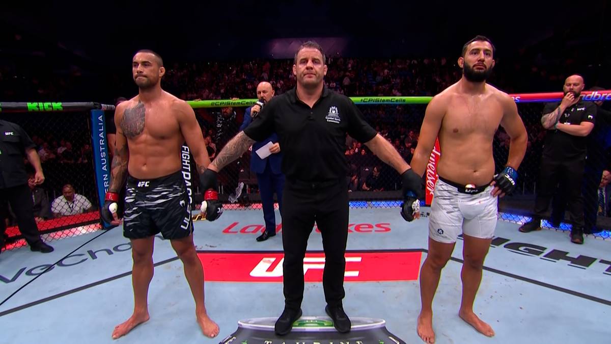 Trzech mężczyzn stoi w ringu UFC. Po lewej i prawej stronie stoją dwaj zawodnicy MMA, a pośrodku sędzia. Wszyscy mają na sobie odzież sportową.