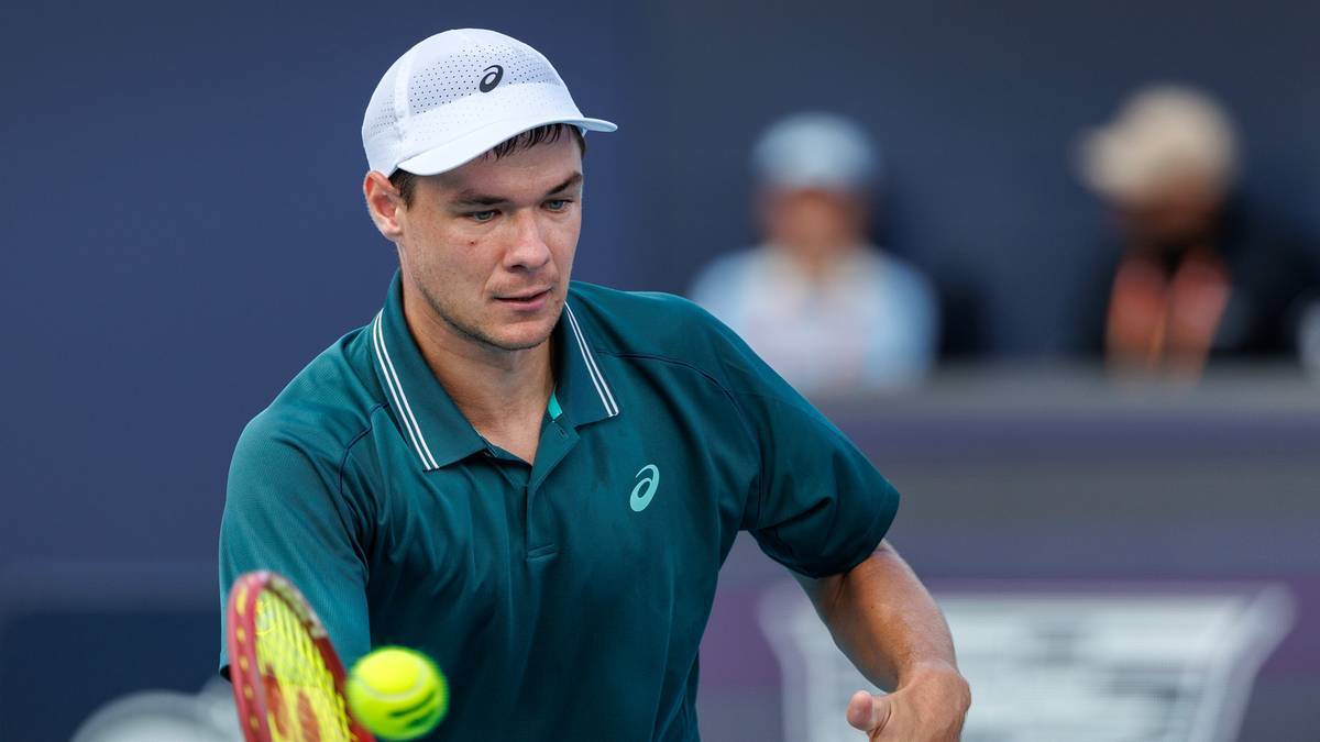 Tenisista w zielonej koszulce i białej czapce uderza piłkę rakietą.