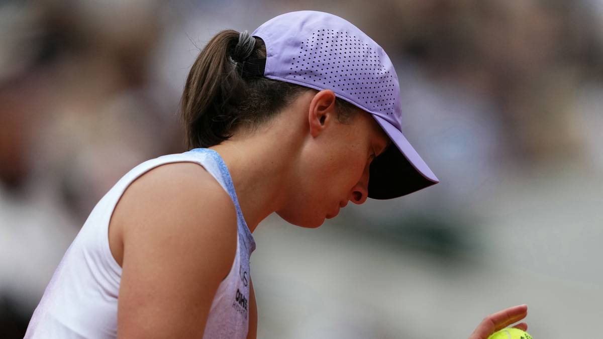 Iga Świątek w czapce z daszkiem i białej koszulce, trzyma w dłoni piłeczkę tenisową, jej głowa jest pochylona w dół.
