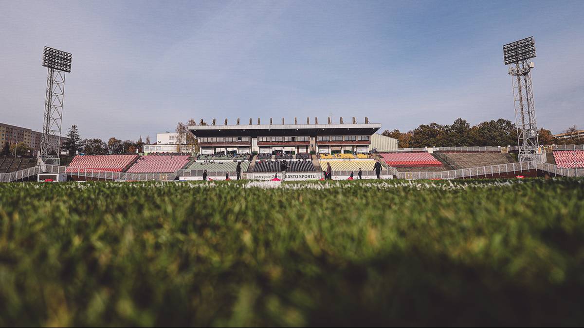 Widok na stadion piłkarski z murawą na pierwszym planie i pustymi trybunami w tle.