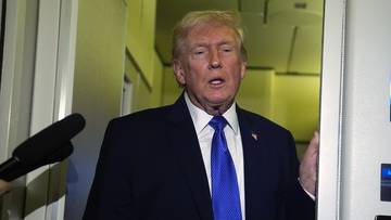 Donald Trump w ciemnym garniturze i niebieskim krawacie, otoczony przez mikrofony.