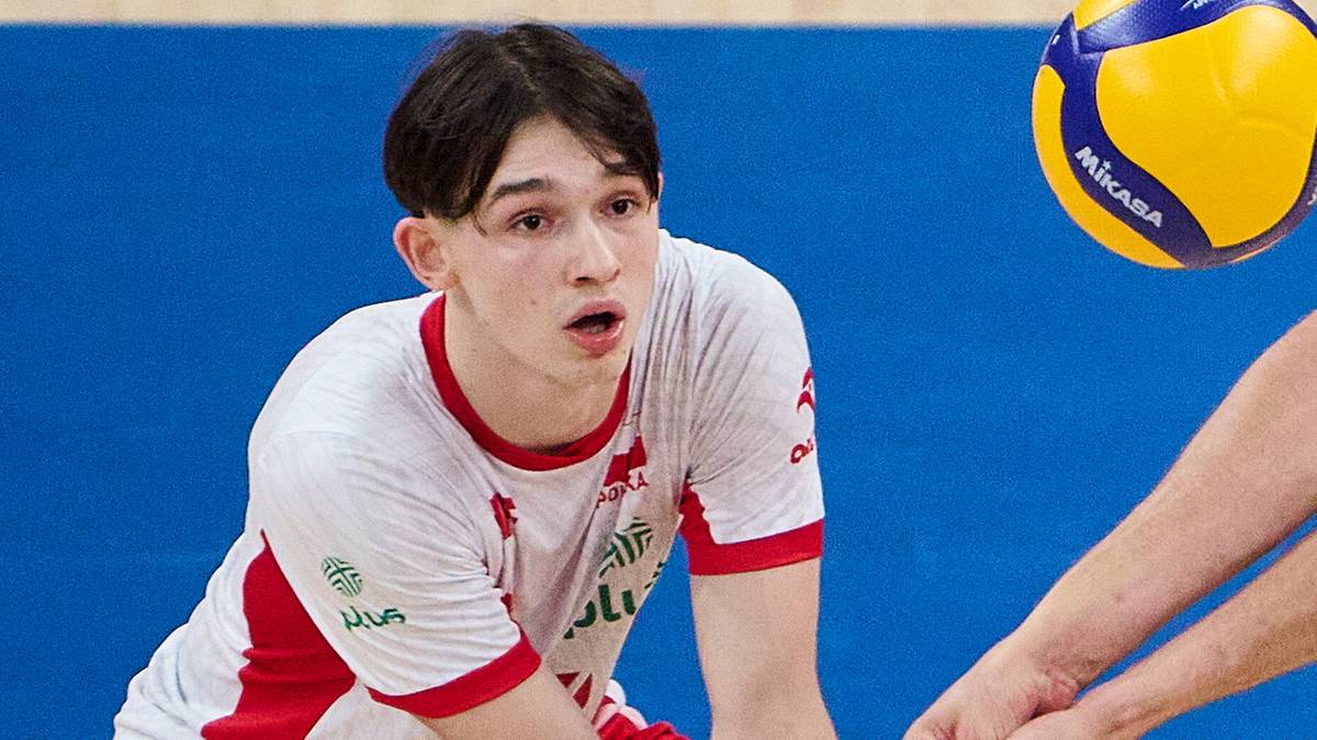 Gracz w siatkówkę w białej koszulce z czerwonymi rękawami, skupiony na piłce, która leci w jego kierunku. W tle niebieskie tło.