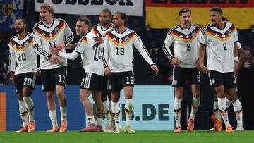 Piłkarze reprezentacji Niemiec w białych strojach z czarnymi wstawkami na zielonym boisku, świętujący razem, z banerami w tle.