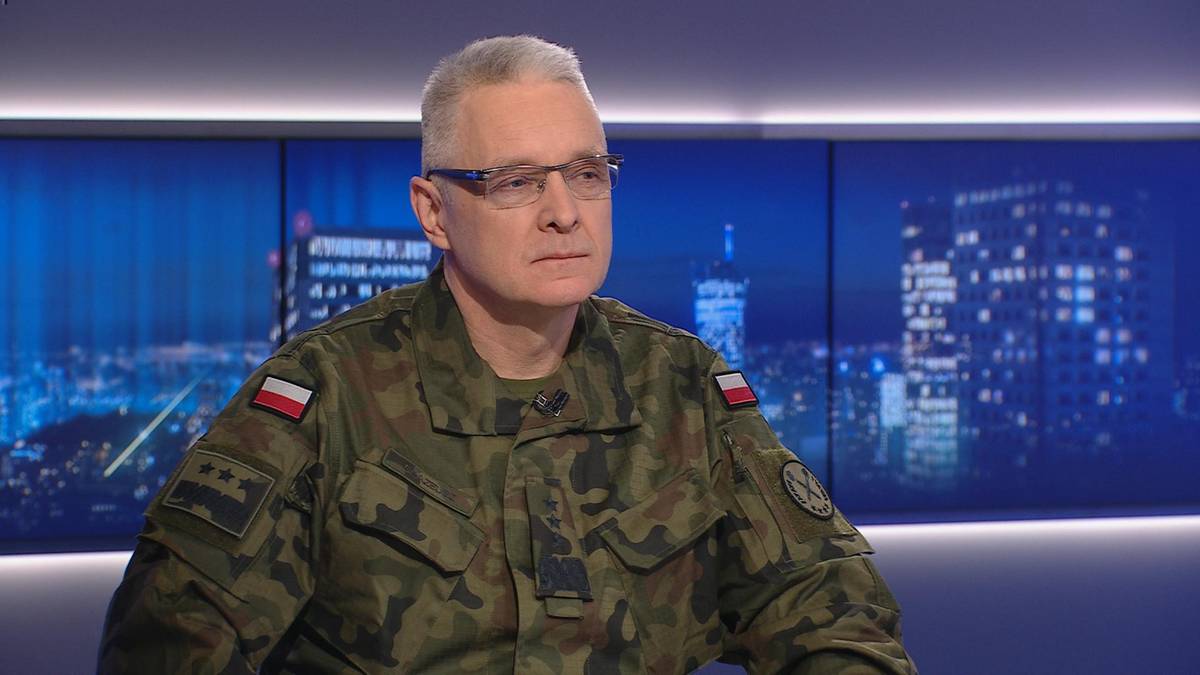 Generał broni Piotr Błazeusz w mundurze wojskowym siedzi przed ścianą z ekranami wyświetlającymi nocne panoramy miast.