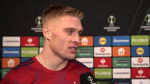 Piłkarz w czerwonej koszulce z kr&oacute;tkim blond włosami i zarostem, udzielający wywiadu na tle ścianki z logotypami sponsor&oacute;w.