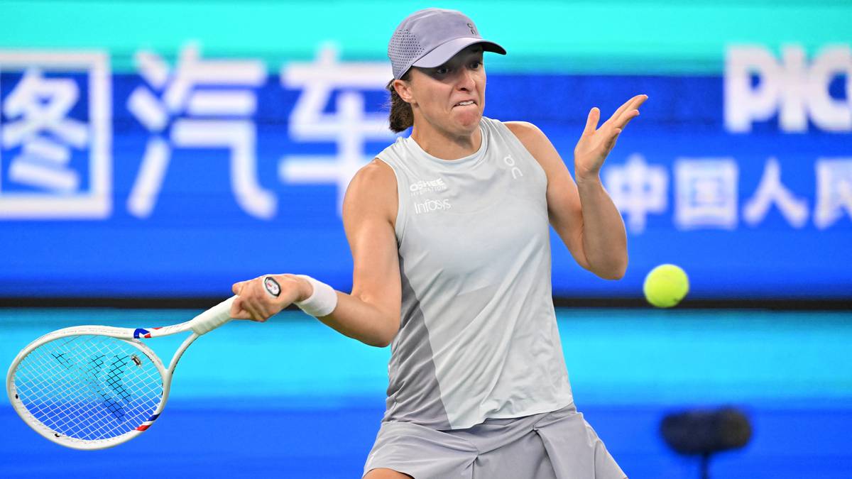 Polka Iga Świątek podczas meczu tenisowego, w białej koszulce i spódniczce, z rakietą w ręku i zmrużonymi oczami, na tle niebiesko-zielonej reklamy.