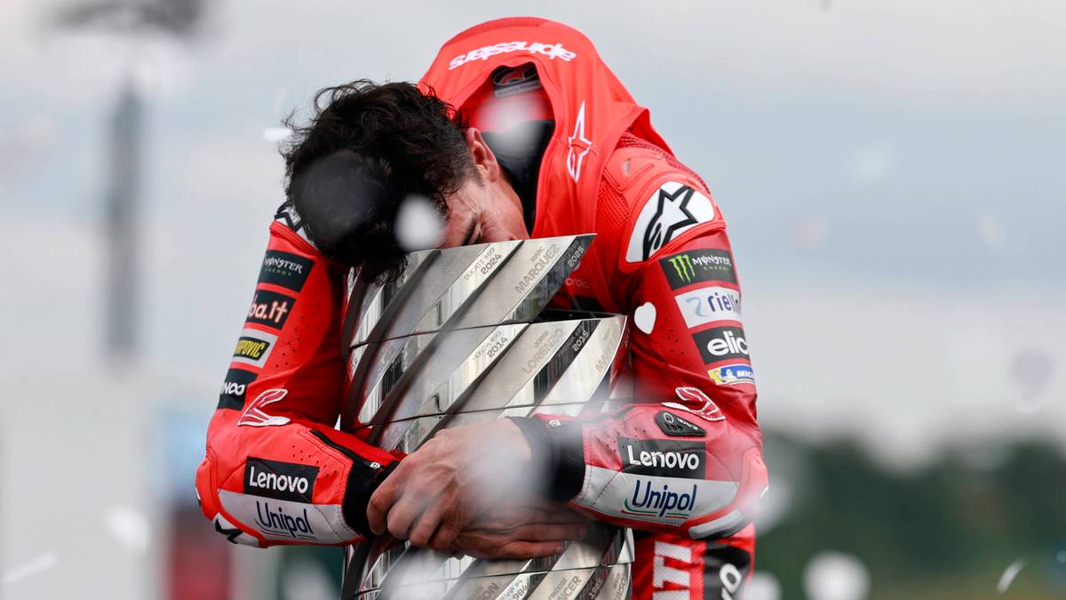 Motocyklista w czerwonym kombinezonie z kapturem obejmuje puchar, wyrażając silne emocje.