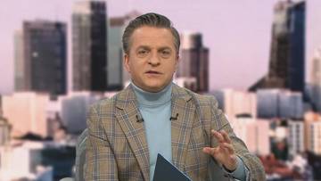 Mężczyzna w marynarce w kratę i niebieskim golfie m&oacute;wi do kamery, gestykulując prawą ręką. W tle widoczne są rozmyte budynki miejskie.