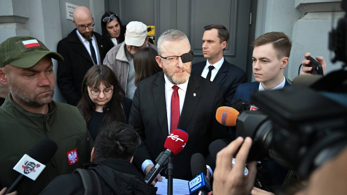 Grupa osób zgromadzona wokół mężczyzny z opaską na oku, trzymającego mikrofon, otoczona przez dziennikarzy.