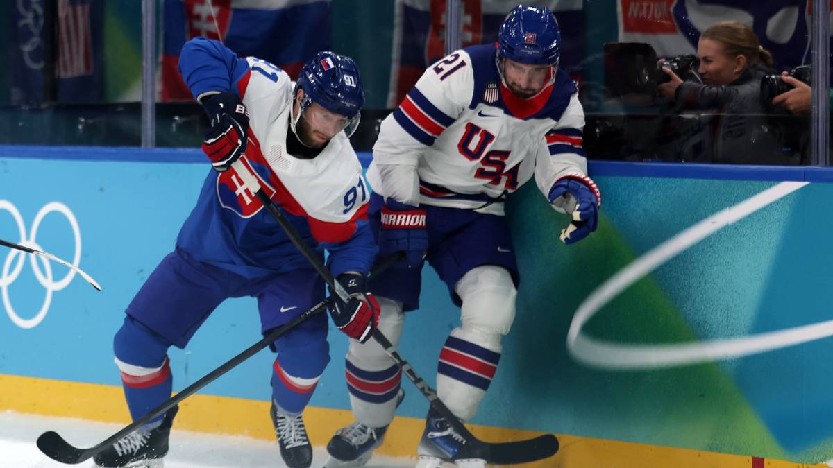 Dw&oacute;ch hokeist&oacute;w walczących o krążek na lodowisku. Jeden zawodnik ubrany jest w niebieski str&oacute;j z numerem 91 i flagą Słowacji, drugi w biały str&oacute;j z napisem USA. Na bandzie w tle widoczne są olimpijskie k&oacute;łka.