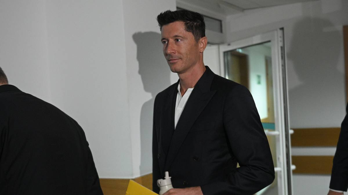 Robert Lewandowski w czarnym garniturze i białej koszuli, trzymający żółty dokument, stoi w korytarzu.