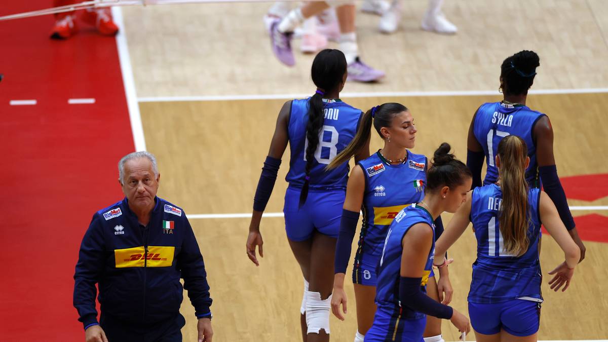 Grupa zawodniczek siatk&oacute;wki w niebieskich strojach stoi na boisku. W tle widać trenera siatkarskiego.