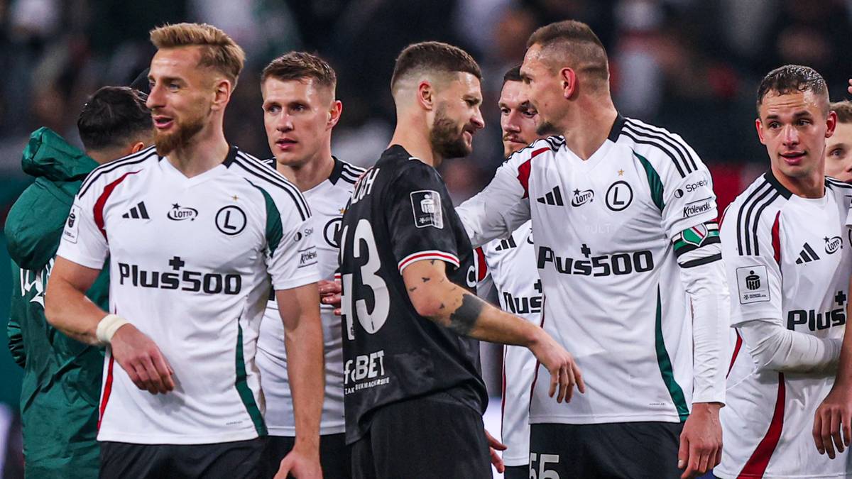 Piłkarze na boisku piłkarskim w białych strojach, niekt&oacute;rzy rozmawiają ze sobą, inni patrzą w stronę publiczności.