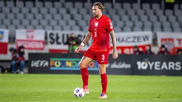 Piłkarz w czerwonej koszulce reprezentacji Polski stoi na boisku z piłką przy nodze.