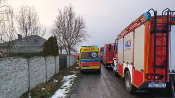 Samochody strażackie i karetka pogotowia stoją na drodze w pobliżu dom&oacute;w.