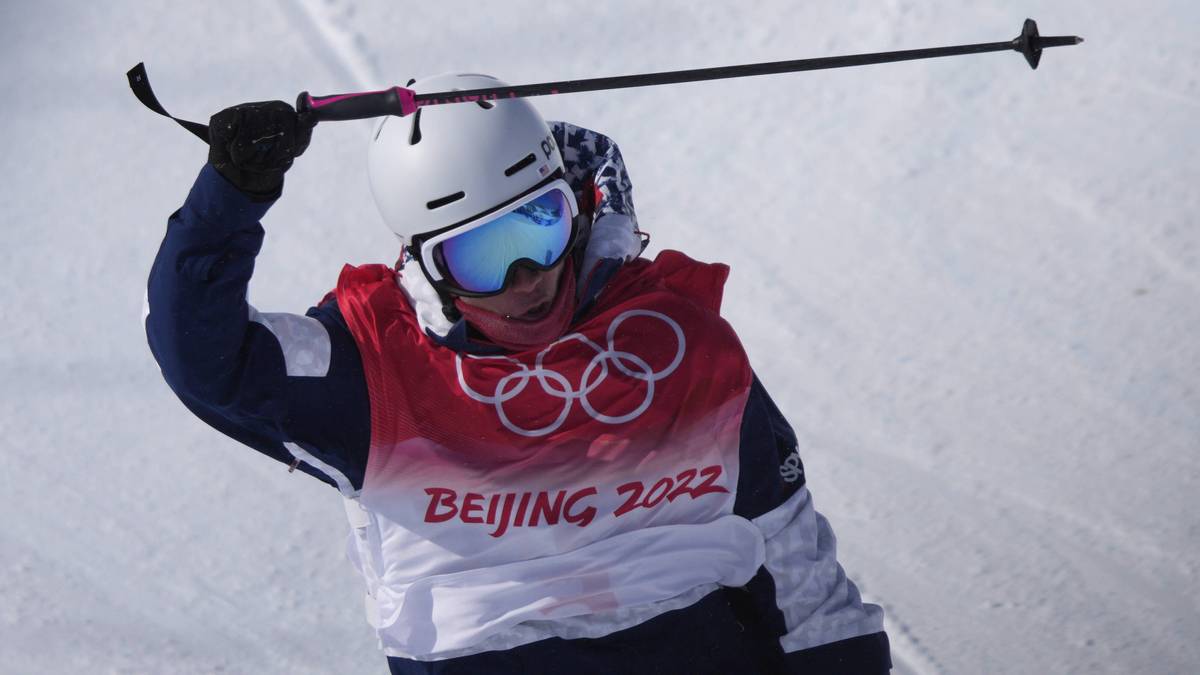 Narciarz w kasku i goglach z kijami narciarskimi trzyma jeden kij nad głową, ubrany w kamizelkę z logo Pekin 2022.