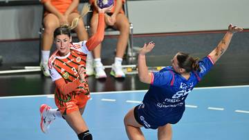 Dwie zawodniczki piłki ręcznej w akcji podczas meczu. Jedna zawodniczka w pomarańczowej koszulce, druga w niebieskiej.