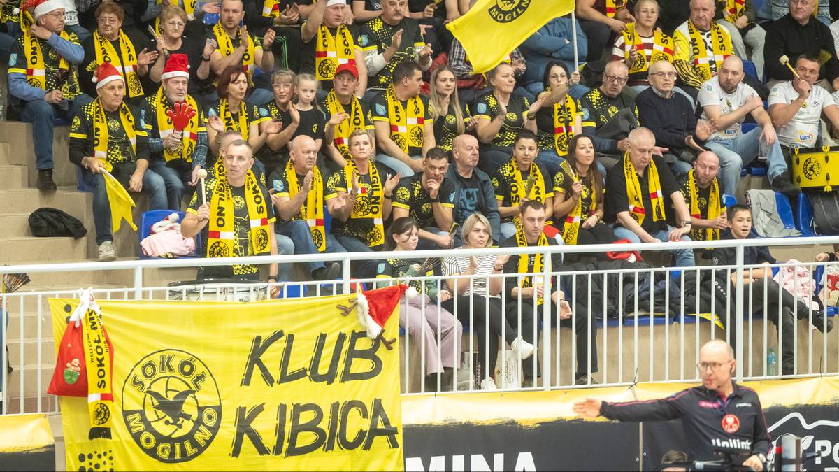 Grupa kibiców w żółto-czarnych strojach siedzi na trybunach, oglądając mecz. Niektórzy mają na głowach czapki mikołaja. Na pierwszym planie widoczny jest żółty transparent z napisem "KLUB KIBICA".