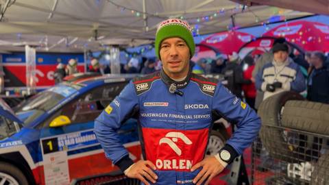 Szymon Gospodarczyk w czapce z pomponem, ubrany w kombinezon rajdowy z logo Orlen i Skoda, stoi przed samochodem rajdowym.