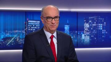 Starszy mężczyzna w okularach, ciemnym garniturze i czerwonym krawacie, uśmiecha się do kamery. W tle widoczne są podświetlone ekrany z panoramą nocnego miasta.