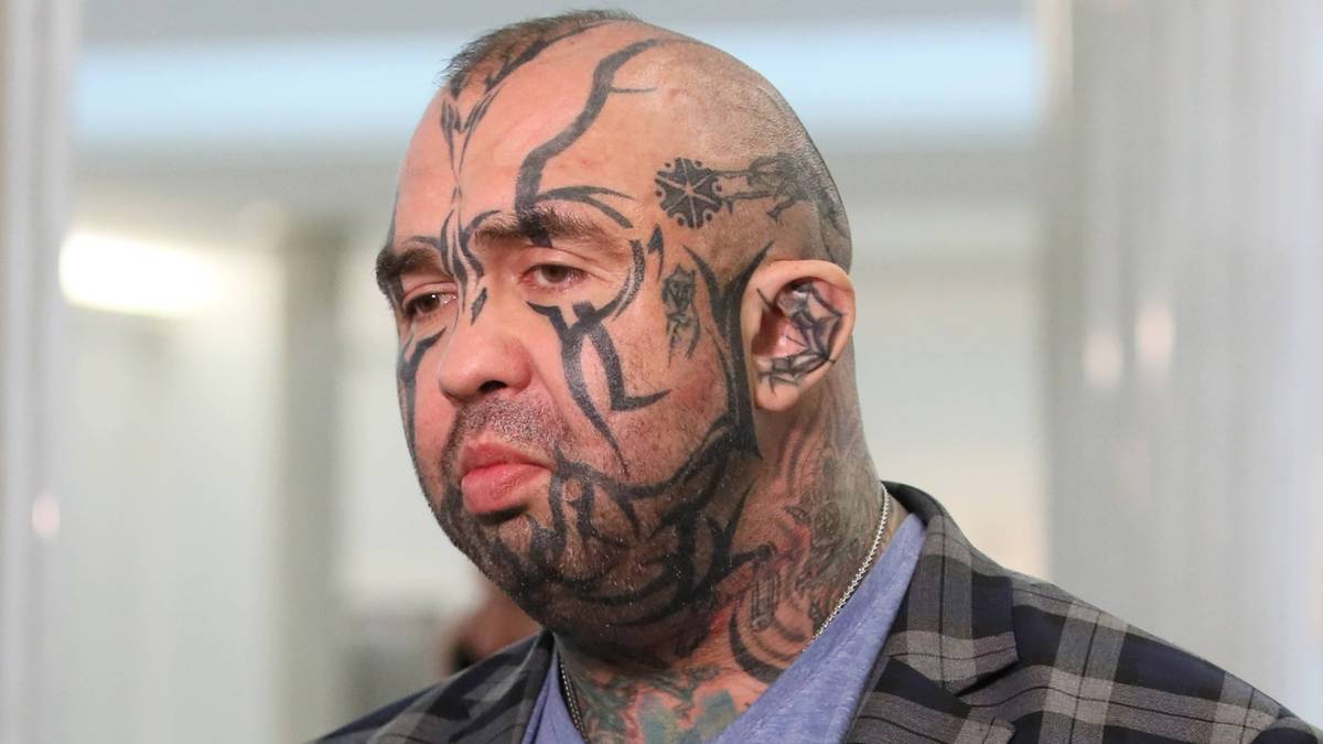 Mężczyzna z licznymi tatuażami na twarzy i szyi udziela wywiadu przed mikrofonem.