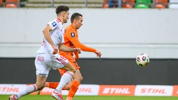 Dw&oacute;ch piłkarzy grających w piłkę nożną, jeden w białym stroju, drugi w pomarańczowym, na boisku piłkarskim.