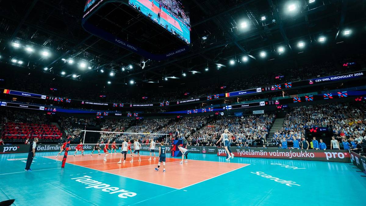Mecz siatk&oacute;wki w hali sportowej z zawodnikami na boisku, siatką i publicznością na trybunach.