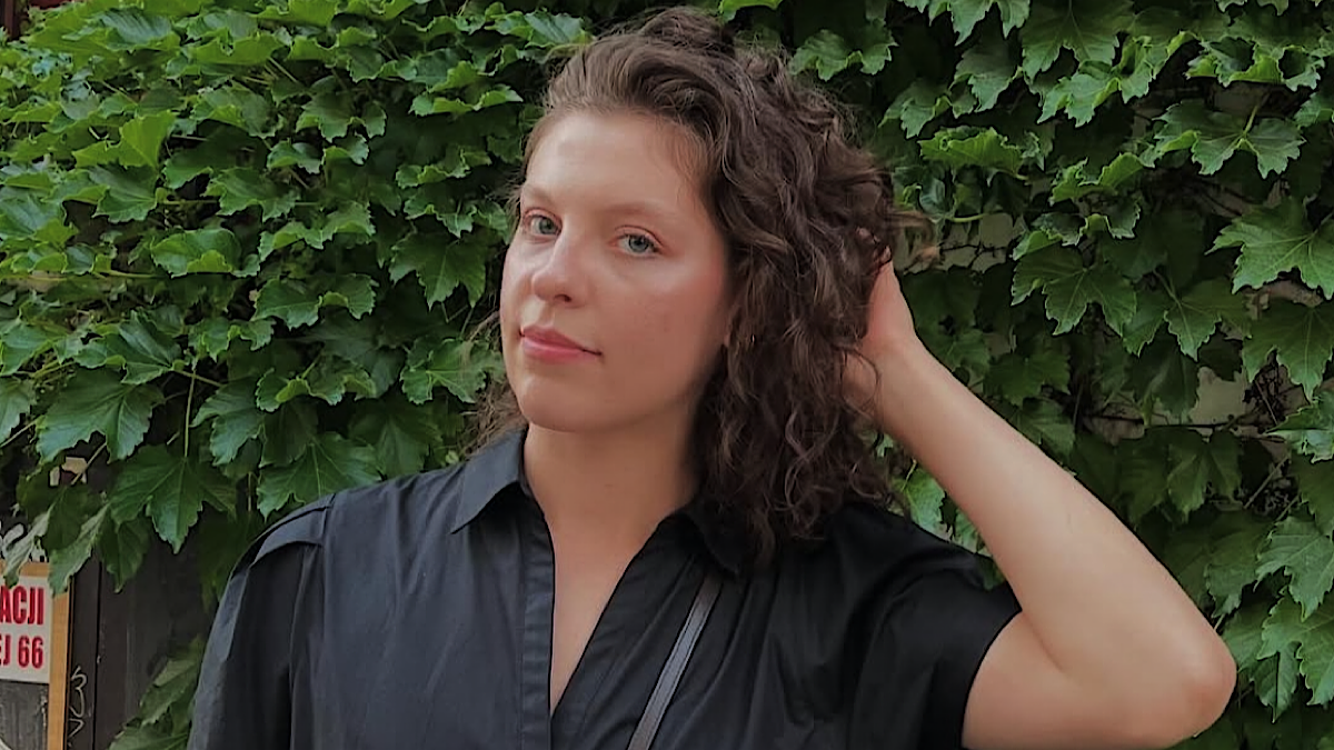 Młoda kobieta o kręconych, ciemnych włosach, ubrana w czarną koszulę, patrzy w obiektyw na tle zielonych liści.