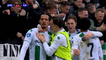 Piłkarze FC Groningen w białych strojach z zielonymi pasami celebrują bramkę, przytulając się na boisku. W tle widać kibic&oacute;w i wynik meczu z informacją o starciu Groningen z Ajaxem.
