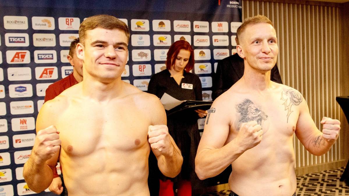 Dwóch mężczyzn w pozycji bokserskiej, z zaciśniętymi pięściami, stojących obok siebie na tle ściany z logo wydarzenia.