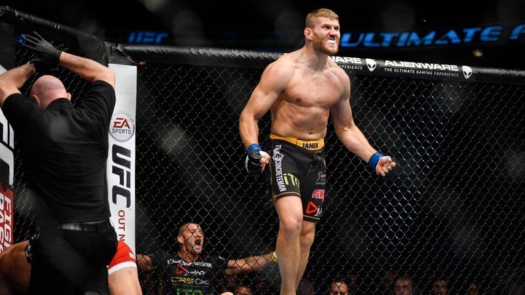 Zawodnik Jan Błachowicz triumfuje po zwycięskiej walce w oktagonie UFC, podczas gdy sędzia przerywa pojedynek i klęczy przy pokonanym przeciwniku.
