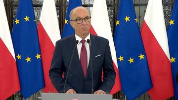 Mężczyzna w garniturze i okularach stoi przy mównicy na tle flag Polski i Unii Europejskiej.