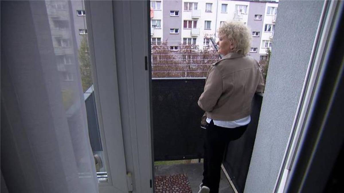 Kobieta o kręconych blond włosach stoi na balkonie, patrząc w dal na osiedle mieszkaniowe.
