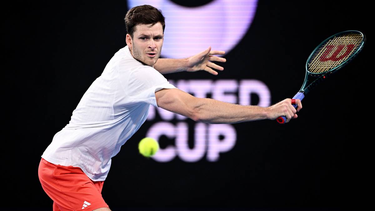 Mężczyzna w białej koszulce i czerwonych spodenkach odbija piłkę tenisową rakietą podczas meczu.