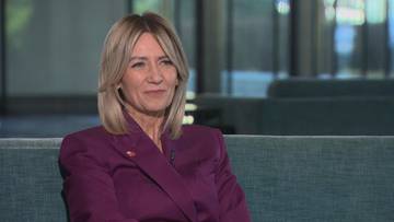 Kobieta w bordowym żakiecie siedzi na kanapie, patrząc lekko w bok z uśmiechem.