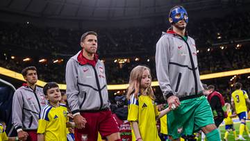 Piłkarze w szarych bluzach wychodzą na murawę stadionu, prowadząc dzieci.