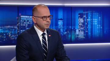 Polityk w okularach i ciemnym garniturze wypowiada się na tle ekranu z widokiem nocnego miasta.