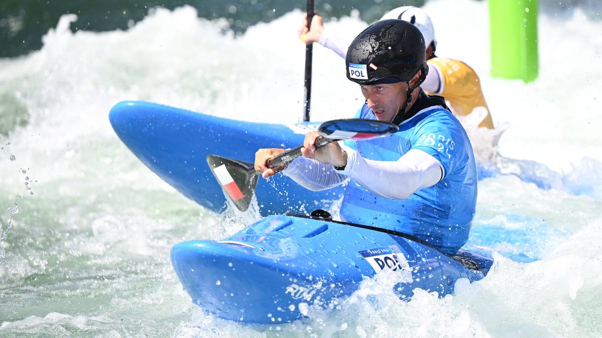 Zawodnik w kasku i okularach pływa kajakiem po wzburzonej wodzie. Ubrany jest w niebieską koszulkę z nadrukiem.