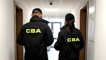 Dwie osoby w czarnych kurtkach z napisem "CBA" na plecach idą korytarzem.