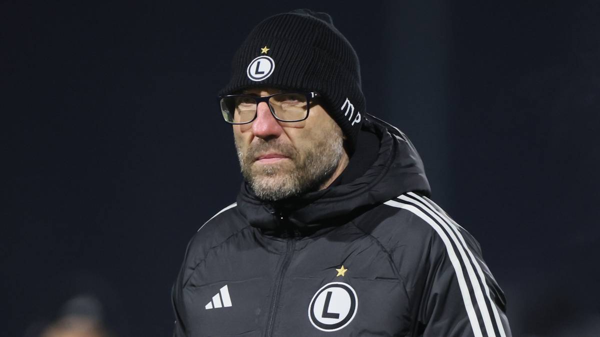 Trener w zimowej kurtce i czapce stoi na boisku.