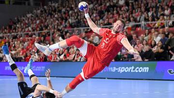 Piłkarz ręczny w czerwonym stroju wykonuje rzut do bramki, drugi zawodnik leży na parkiecie.