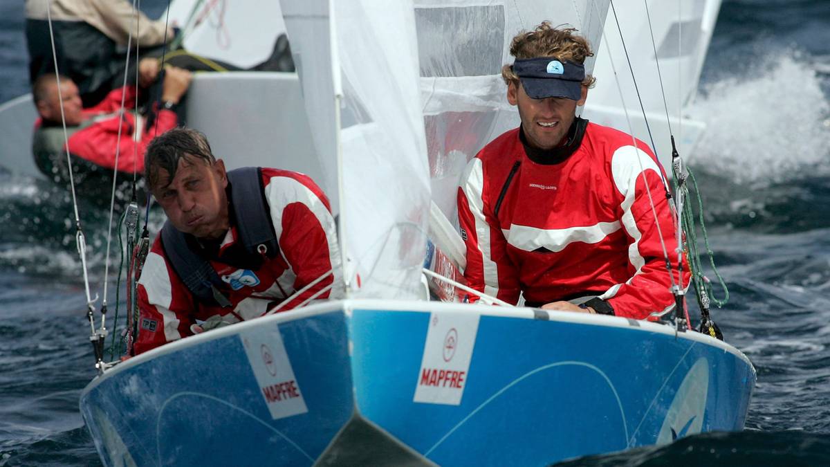 Dwóch mężczyzn w czerwono-białych bluzach żeglarskich siedzi w niebieskiej łodzi żaglowej na wzburzonej wodzie, z białymi żaglami w tle.