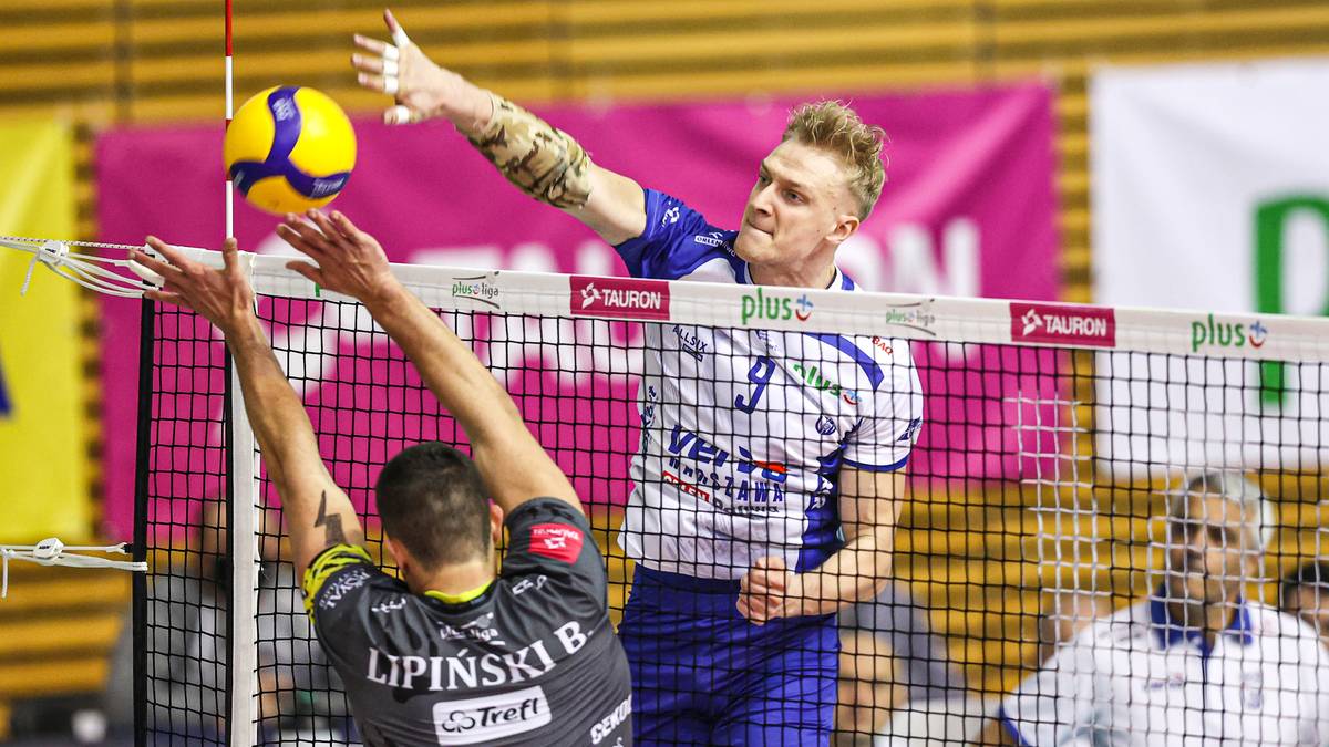 Siatkarz w białej koszulce z numerem 9 i niebieskimi wstawkami wykonuje atak, zagrywając piłkę nad siatką. Naprzeciwko niego, drugi siatkarz w czarnej koszulce z numerem 1 próbuje zablokować jego zagranie. W tle widoczni są inni zawodnicy oraz fragment trybun.