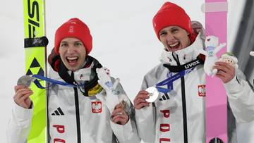 Dw&oacute;ch polskich skoczk&oacute;w narciarskich w strojach z biało-czerwonymi elementami i czerwonymi czapkami pokazuje medale, stojąc obok nart.