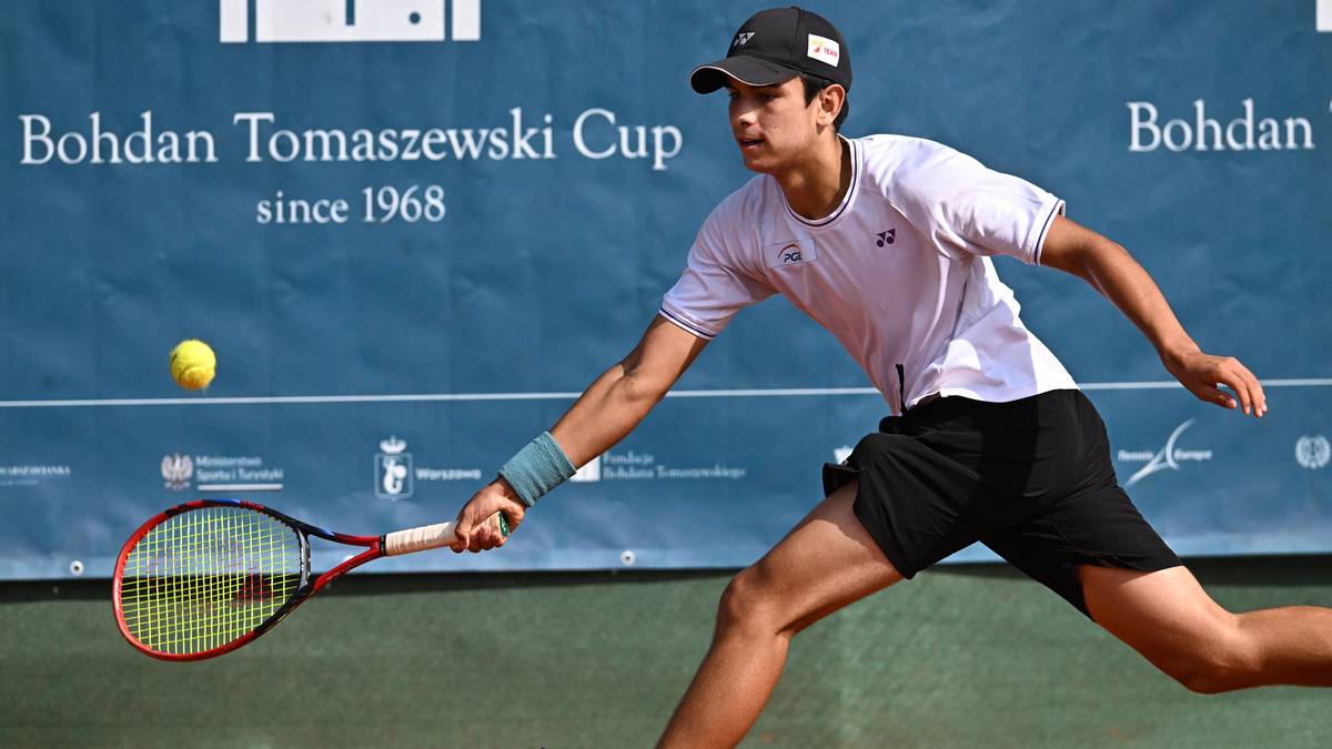 Młody mężczyzna w białej koszulce i czarnej czapce z daszkiem gra w tenisa, wykonując uderzenie po swoim lewym boku.