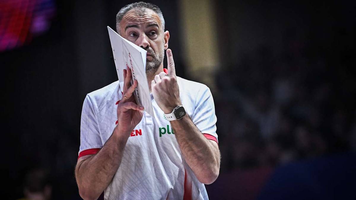 Trener siatkówki w białej koszulce z logotypami "PL" i "EN", trzymający kartkę z notatkami i pokazujący palcem do góry.