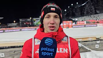 Kacper Tomasiak, polski skoczek narciarski, w czapce i kurtce z logo "Polsat Sport", na tle skoczni narciarskiej.