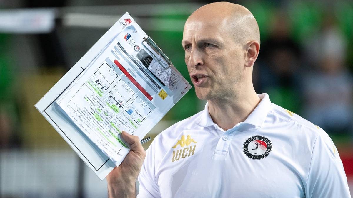 Trener siatkówki w białej koszulce z kołnierzykiem, trzymający w prawej dłoni dokument z rozpiską taktyczną. Ma łysą głowę i skupiony wyraz twarzy.